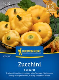 Gelbe, runde Zucchini mit Stiel in einem schwarzen Korb, umgeben von grünen Blättern und gelben Blüten.