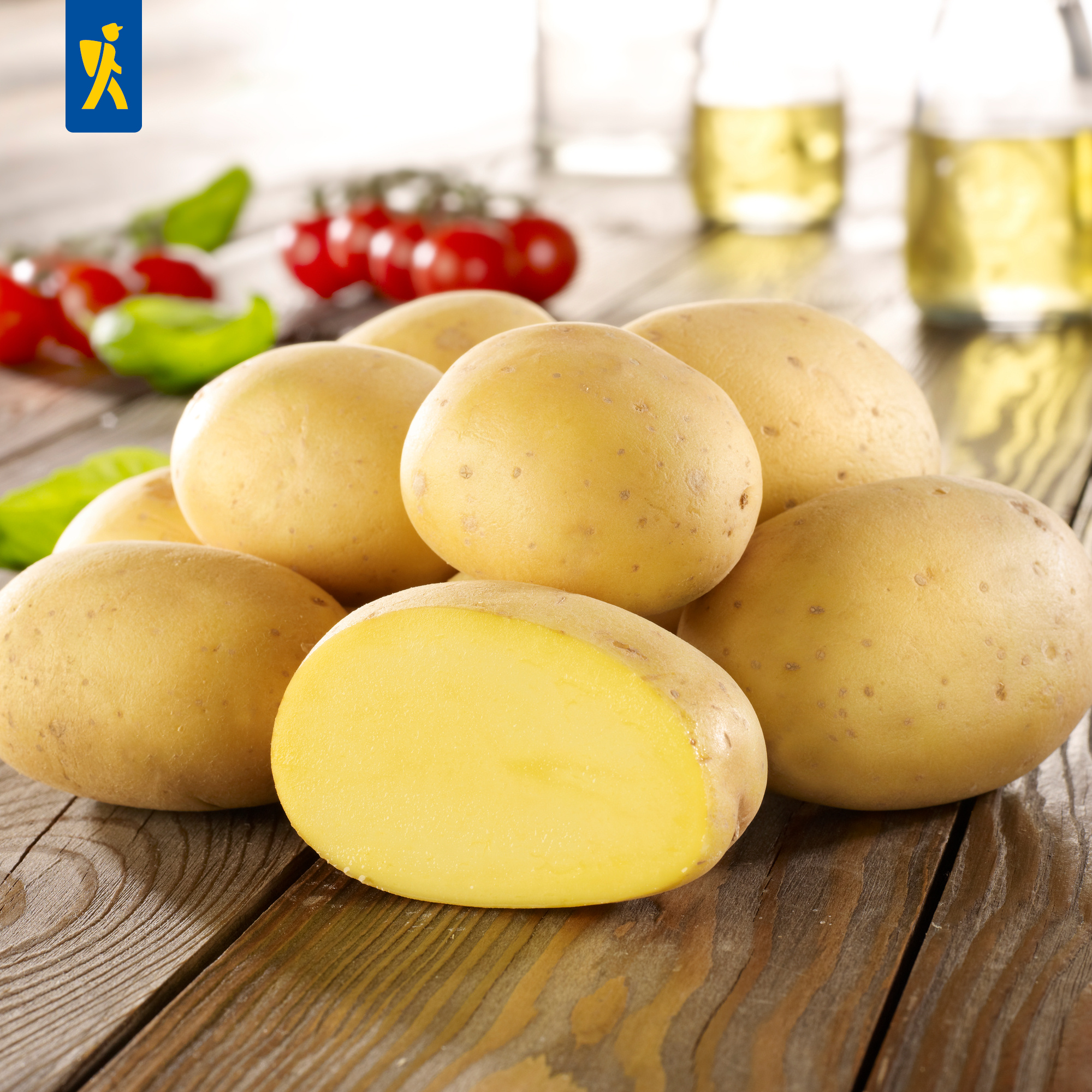 Mehrere rohe gelbe Kartoffeln auf einem Holzbrett, im Hintergrund Tomaten, Basilikum und Gläser mit einem Getränk.