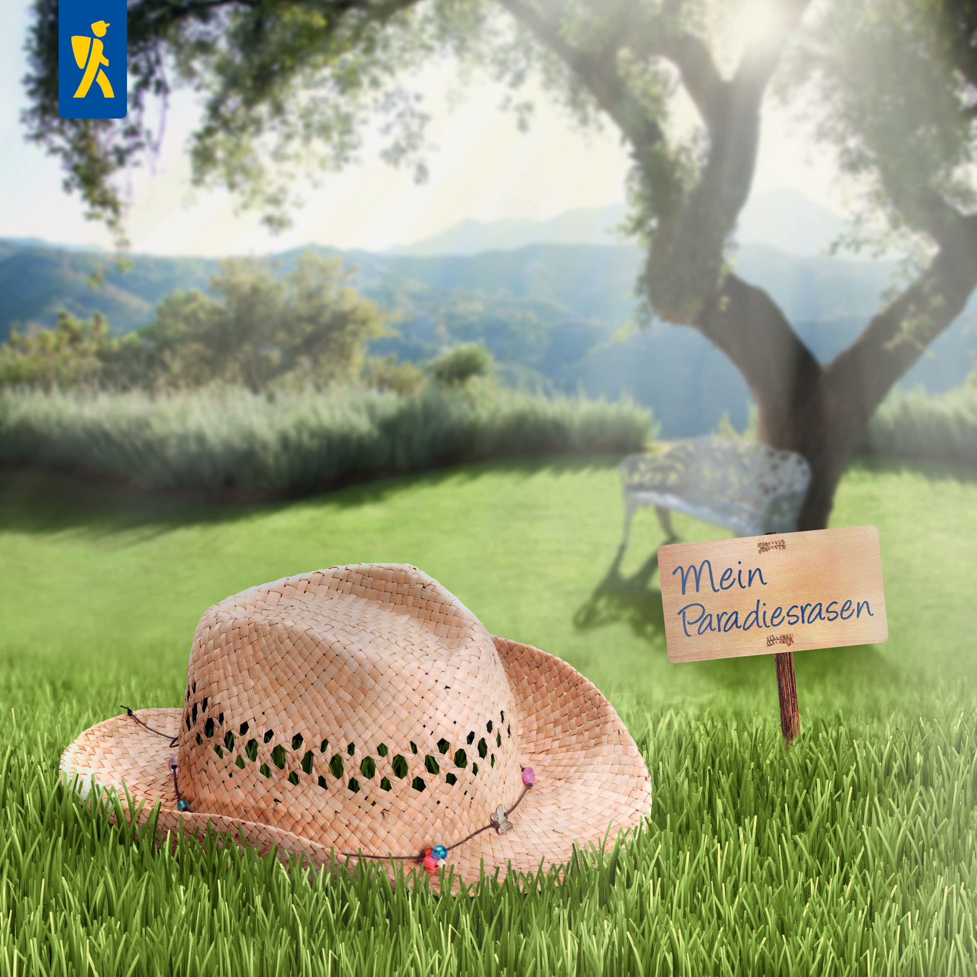 Sonnenhut liegt auf Gras, Holzschild mit "Mein Paradiesrasen", Baum, Wiese, Bank und Berge im Hintergrund.