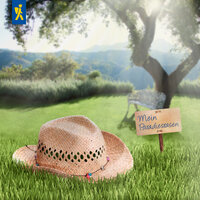 Sonnenhut liegt auf Gras, Holzschild mit "Mein Paradiesrasen", Baum, Wiese, Bank und Berge im Hintergrund.