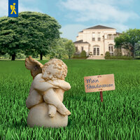 Kleiner geschnitzter Engel mit gefalteten Händen und Flügeln auf grünem Rasen vor großem weißen Haus.